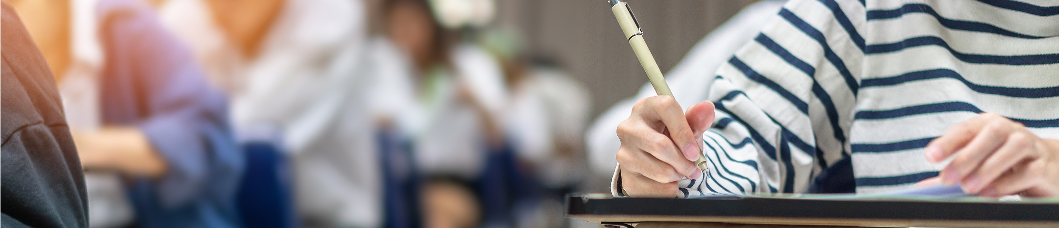 Eine Person sitzt an einem Tisch und schreibt mit einem Stift auf Papier, im Hintergrund sind weitere Personen zu sehen.