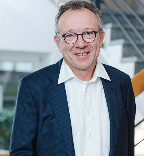 Lächelnder Mann mit Brille in einem hellen Raum mit Treppe im Hintergrund. Er trägt einen Anzug und ein weißes Hemd.