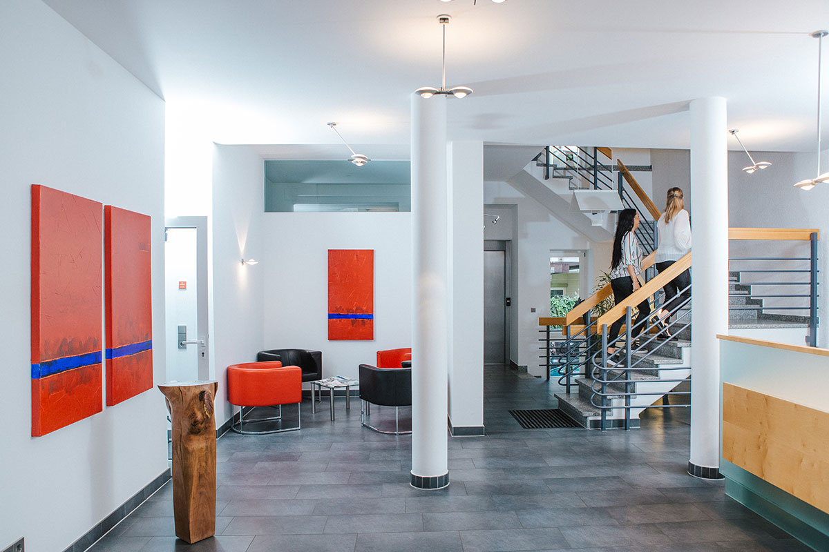 Moderne Empfangshalle mit roten Kunstwerken, bequemen Sesseln und Treppe im Hintergrund. Zwei Personen gehen die Treppe hoch.