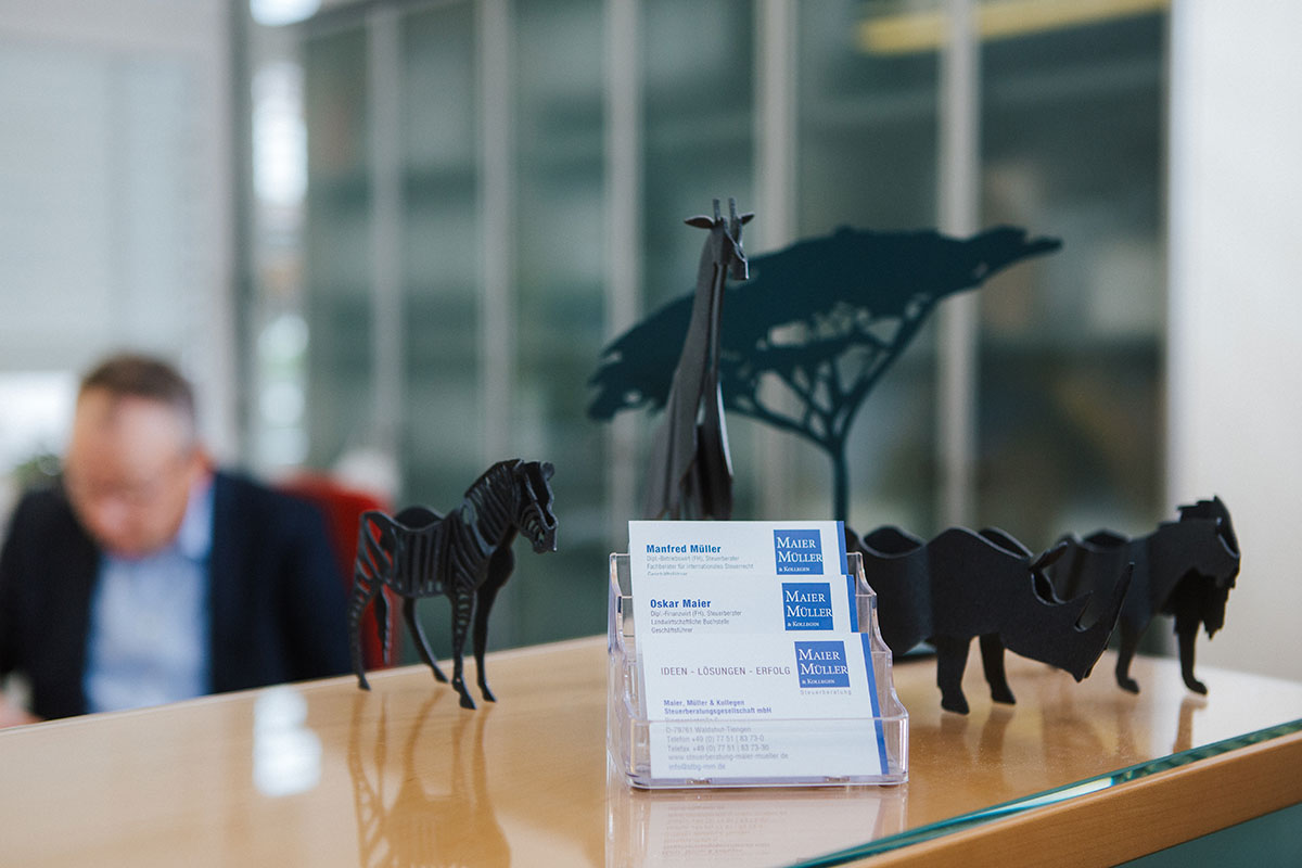 Schreibtisch mit Visitenkartenhalter, umgeben von stilisierten Tierfiguren und einer verschwommenen Person im Hintergrund.