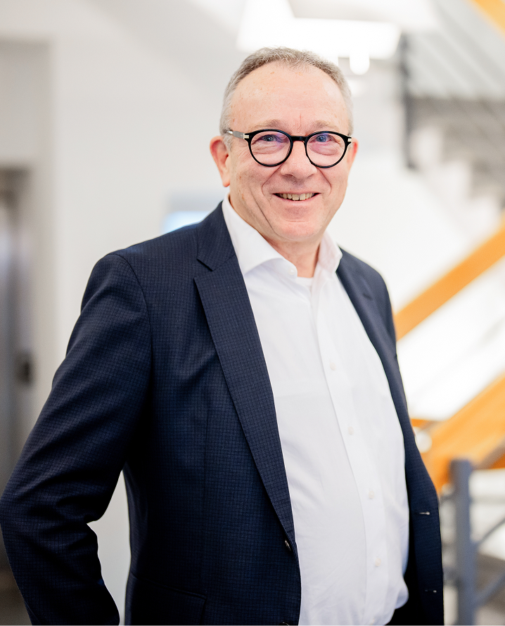 Ein lächelnder Mann mit Brille steht in einem modernen Innenraum, trägt Anzug und schaut freundlich in die Kamera.