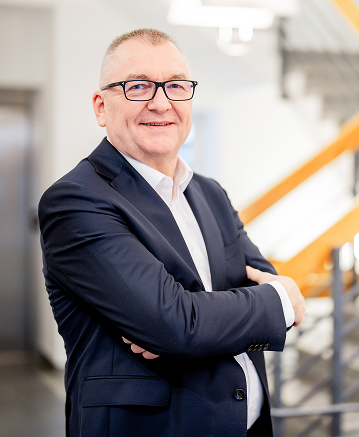 Mann mit Brille in Anzug, lächelnd, vor einer Treppe in einem modernen Raum.