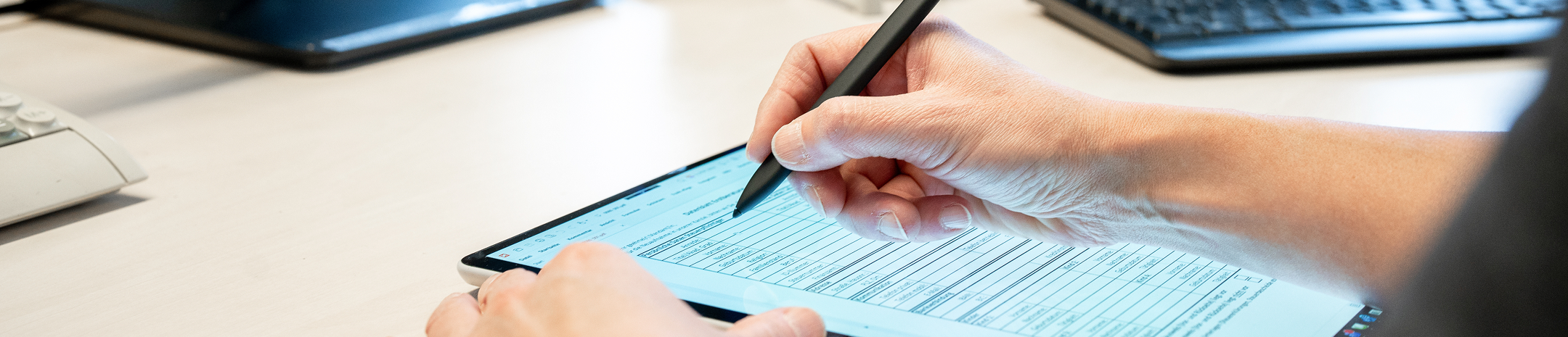 Eine Person schreibt mit einem Stylus auf einem Tablet, während ein Computer im Hintergrund sichtbar ist.